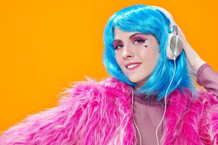 Attractive party girl with bright pink makeup and blue wig wearing pink fur coat and headphones on a yellow background. Make-up and cosmetics, hairstyle. Fashion girl. Copy space.の写真素材