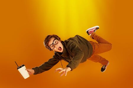Emotional boy teenager in glasses flies with a glass of coffee and screams in surprise. Yellow background. Copy space. の写真素材