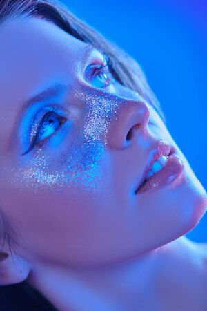 Party and holiday style. Close-up portrait of a beautiful girl with shiny glitter freckles posing in blue and pink light. Cosmetics and makeup.の写真素材