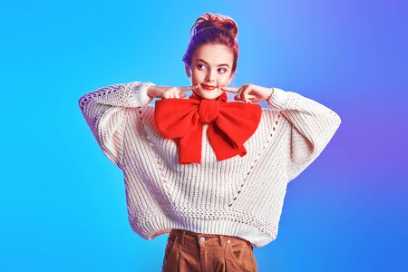 Portrait of an excited pretty girl in a white sweater and with a big elegant red bow on her neck against a soft pink and blue light. Beauty, fashion concept. Winter style.の写真素材