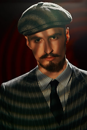 Portrait of a handsome man in elegant classical suit and a cap standing in the dark, illuminated by...