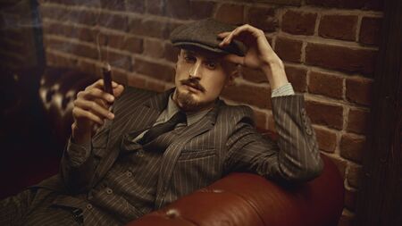Portrait of a handsome courageous man sitting pensive in a rich setting on a leather sofa and smoking a cigar. Luxurious lifestyle. Retro style. Mafia and criminal world, detective.の写真素材