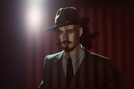 Portrait of a handsome man in elegant classical suit and a hat standing in a dark room lit by...