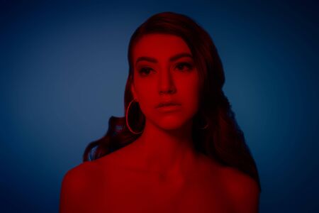 Art portrait. Beautiful young woman with dark wavy hair posing in red light on a dark blue background. Beauty and fashion concept.の写真素材
