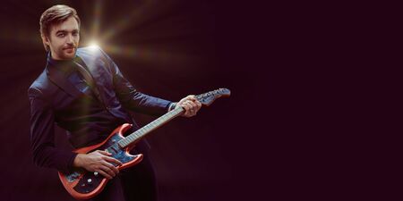 Emotional cheerful young man musician in classic suit plays rock'n'roll on electric guitar. Dark background with copy space.の写真素材