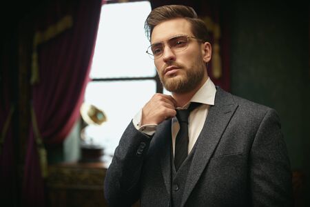 Handsome gentleman poses in a tweed suit and glasses and adjusts his shirt collar in a vintage interior. Luxurious lifestyle. Retro style.の写真素材
