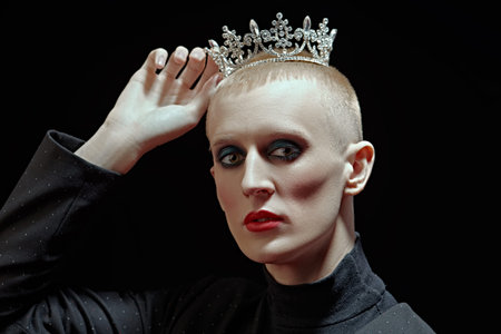 Portrait of a young man with women’s makeup and with a crown on his head on a black background....