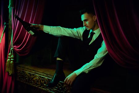 A young guy in a classic suit is sitting on the windowsill with a bottle of wine in a vintage interior. Lifestyle of rich people.の写真素材