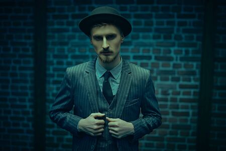 Portrait of a handsome young man in elegant suit and bowler hat standing in blue light by a brick wall. Men's fashion. Luxurious lifestyle.の写真素材