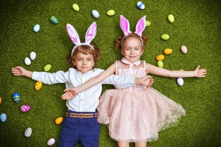Cute smiling boy and girl with bunny ears are lying on the grass with colorful eggs. Easter holiday.の写真素材
