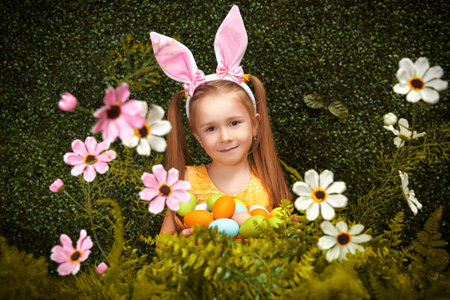 Cute smiling girl with bunny ears is holding holding a basket with flowers and colorful eggs surrounded by greenery and flowers. Spring-summer session. Easter holiday.の写真素材