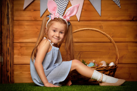 Easter holiday. Happy little girl with bunny ears is posing with basket of colorful eggs  on a grass...