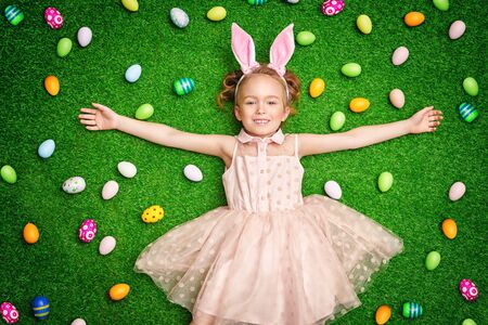 Funny little girl with bunny ears lying on a grass among colorful Easter eggs. Easter holiday.の写真素材