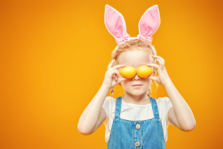 Funny little girl is posing in light t-shirt and sundress is holding Easter painted eggs on a yellow...