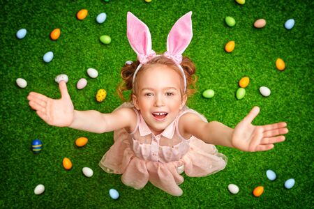 Cute little girl with bunny ears are posing with her hands raised on grass background with colorful eggs. Easter holiday.の写真素材