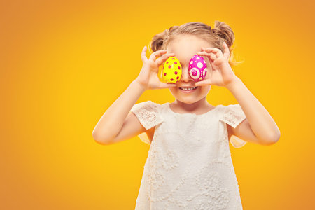 Funny little girl is posing in light dress holding Easter painted eggs on a yellow background in the...