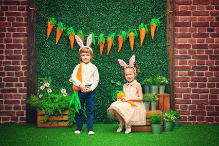 Cute smiling boy and girl with bunny ears are holding carrots on the background of Easter...