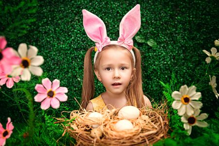 Cute smiling girl with bunny ears holds a nest with eggs in a spring garden. Easter holiday.の写真素材