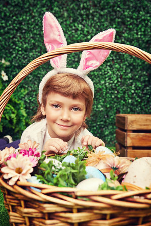 Happy little boy with bunny ears sits with a basket of easter eggs on a green lawn in a spring...