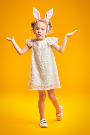 Easter holiday. Cute little girl poses in light dress and with bunny ears on a yellow background. Studio portrait. Children's summer and spring fashion.の写真素材