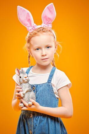 Cute red-haired girl holds toy Easter bunny and smiles. Happy childhood. Easter holiday. Studio portrait on a yellow background.の写真素材