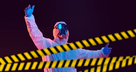 COVID-19 concept. A medicine worker in a protective suit stands behind restrictive tapes and makes a warning gesture with his hands. Isolation and quarantine.の写真素材