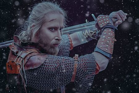Portrait of a brave medieval warrior in armor with a sword on a black background with snowfall. The Witcher man.の写真素材