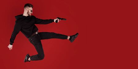 Handsome man in black clothes in motion in flight with a gun. Super agent on a mission. Criminal world, counterterrorism. Studio shot on a red background.の写真素材