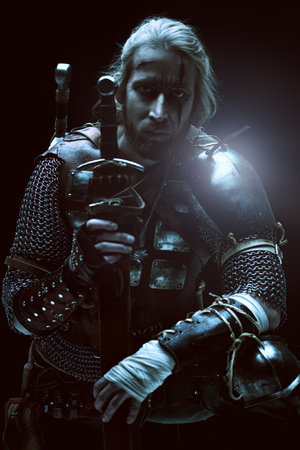 Portrait of a brave medieval warrior in armor with a sword on a black background. The Witcher man.
