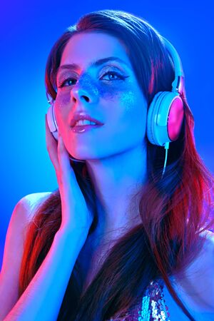 Party and holiday style. Pretty girl with shiny make-up and shiny dress listening to music in headphones and dancing in blue and pink light.の写真素材