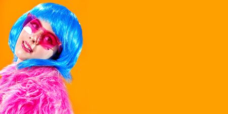 Portrait of an attractive party girl with bright pink makeup and blue wig wearing pink fur coat and sunglasses on a yellow background. Make-up and cosmetics, hairstyle. Fashion girl. Copy space.の写真素材