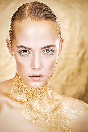 Beauty, fashion gold concept. Portrait of a beautiful girl model with golden skin and golden make-up on a glowing gold background. Jewelry. Cosmetics and beauty care.の写真素材