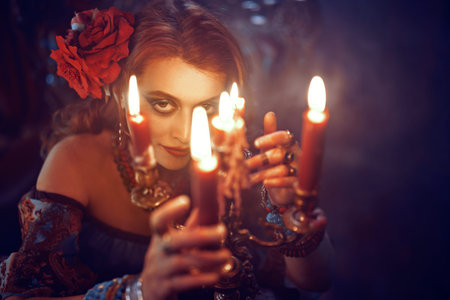 Bewitching woman fortune-teller witch looking at the camera through the flame of candles. Sepia...
