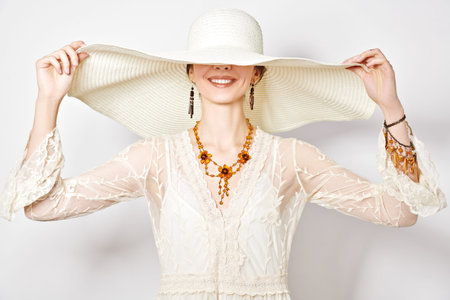 Portrait of a charming young woman in light elegant lace dress and a wide brim hat smiling happily...