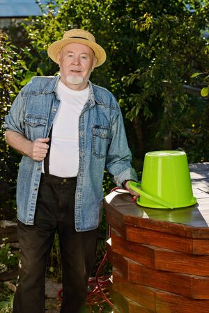 Portrait of an elderly gardener standing with a bucket at the well in the garden. Summer vacation in the country. Work in the garden.の写真素材
