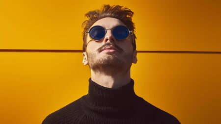 Urban style. Portrait of a stylish young man in black pullover and round mirror sunglasses posing on a street by the yellow industrial wall. Male fashion. Copy space.の写真素材