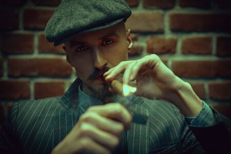 Portrait of a handsome courageous man sitting pensive in the dark and smoking a cigar. Luxurious lifestyle. Retro style. Mafia and criminal world, detective.の写真素材