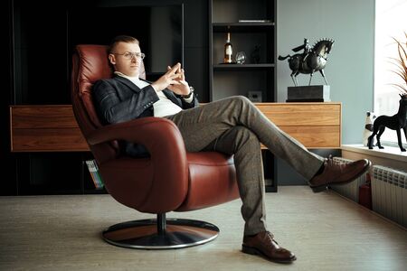 Portrait of a respectable middle-aged businessman in elegant suit and glasses sitting in a leather chair and ponders an important question. Success and leadership.の写真素材