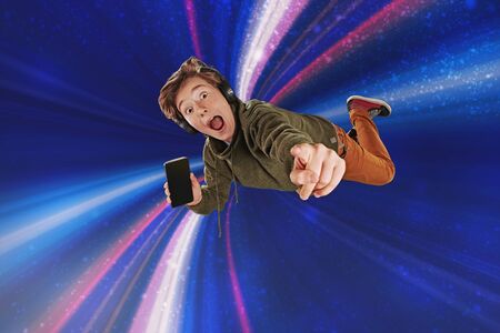 Emotional teen boy is flying through the air like a super man, shouting and showing his smartphone. Dark blue background with colored light beams. Copy space.の写真素材