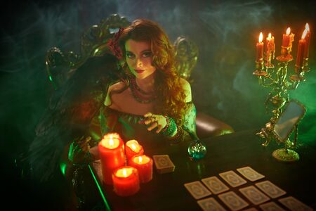 A beautiful red-haired fortune teller is talking to a black raven. The atmosphere of magic and sorcery, magic crystal ball and tarot cards.の写真素材