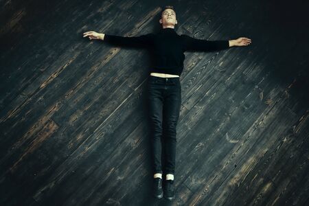 Handsome young man in black clothes lies on a black wooden floor, arms spread out to the sides. Religious concept.の写真素材