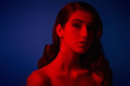 Art portrait. Beautiful young woman with dark wavy hair posing in red light on a dark blue background. Beauty and fashion concept.の写真素材