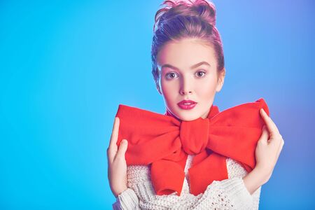 Portrait of an elegant beautiful girl in a white sweater and with a big red bow on her neck against a soft pink and blue light. Beauty, fashion concept. Winter style.の写真素材