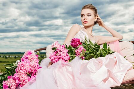 Fashion shot. Gorgeous young woman in a luxuriant pale pink dress lies on the couch among beautiful peony flowers. The backdrop of a landscape with blue sky and white clouds. Wedding style.の写真素材