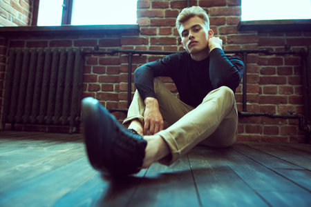 Male beauty, fashion. Portrait of a handsome young man with blond hair sitting on a floor in loft...