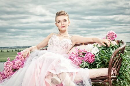 Fashion shot. Gorgeous young woman in a luxuriant pale pink dress lies on the couch among beautiful peony flowers. The backdrop of a landscape with cloudy sky. Wedding style.の写真素材