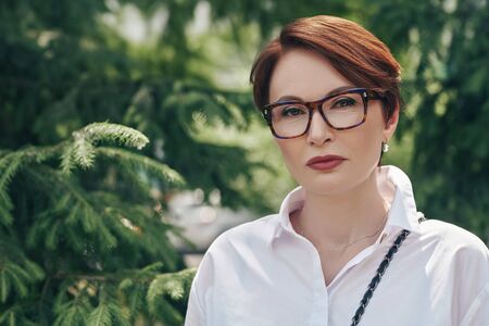 Elegant middle-aged woman in glasses and white shirt stands on a street on a summer day. Business woman portrait. Optics style.の写真素材