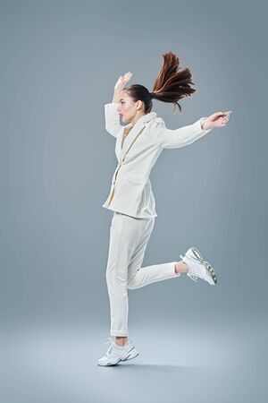 Fashion shot. Portrait of a beautiful young woman in a white suit jumping and running on a grey background.の写真素材