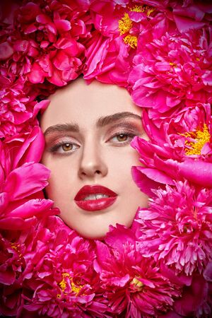 Beauty and flowers. Close-up portrait of a beautiful brunette woman with bright make-up lying among peony flowers. Cosmetics, make-up. Perfumery.の写真素材