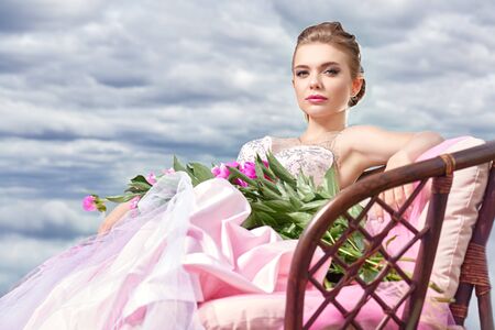 Fashion shot. Gorgeous young woman in a luxuriant pale pink dress lies on the couch among beautiful peony flowers. The backdrop of a landscape with blue sky and white clouds. Wedding style.の写真素材